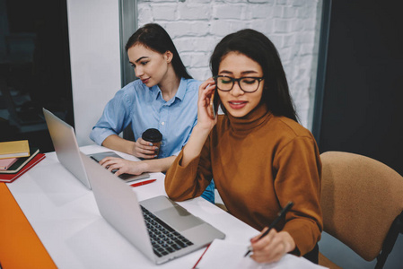 坐在办公室里的年轻妇女集中精力在他们的任务上通过膝上型计算机来拨打电话和监控新闻, 忽略了生产具身的沟通。