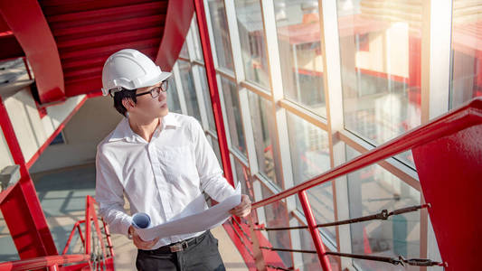 年轻的亚洲男性工程师或建筑师持有蓝图在建筑工地的红色楼梯上佩戴防护安全帽。土木工程建筑与建筑概念
