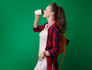 微笑的现代学生妇女在红色衬衣与背包和耳机喝咖啡从白色纸杯子在绿色背景
