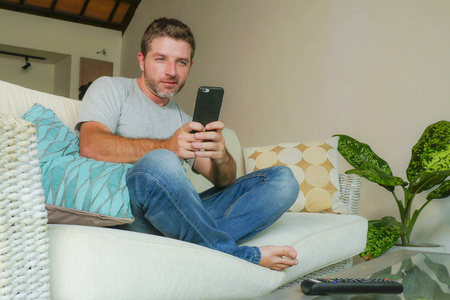 时尚的生活室内肖像年轻英俊和迷人的快乐的人使用互联网交友在线应用程序在手机或网络上坐在家里的沙发沙发与智能手机放松