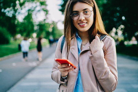 半长肖像欢快的时髦女孩的眼镜花时间在街上的春天一天使用手机, 迷人的年轻女子满意的良好的4g 连接通过智能手机聊天