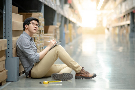 年轻的亚裔男子工人用数码纸和钢笔在仓库货架上的纸板箱里清点产品。物理库存盘点概念