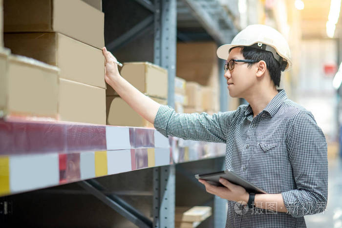 年轻的亚裔男子工人戴安全帽和眼镜在仓库的货架上用数码片和钢笔清点