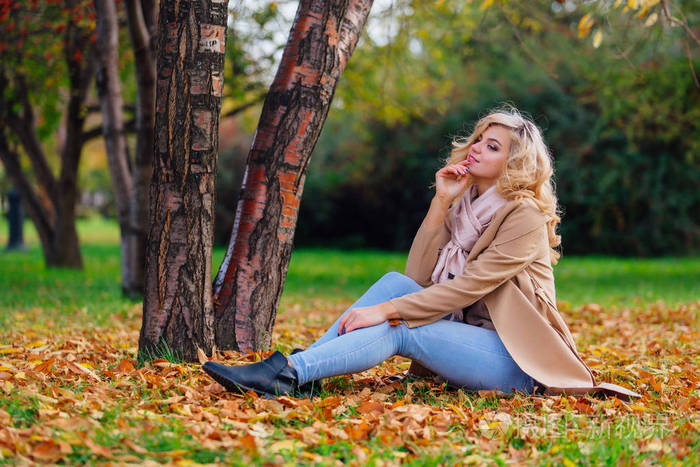 年轻美丽的女人坐在地上的树下覆盖秋天的黄叶