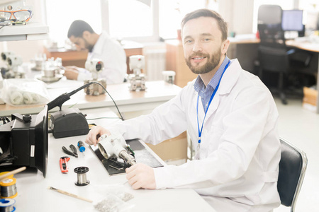 开朗自信的胡子男性修复工程师在实验室大衣坐在桌子上的工具和拆卸测量设备和看相机
