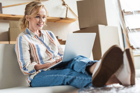 美丽的微笑成熟的女人使用笔记本电脑, 坐在沙发上的新房子