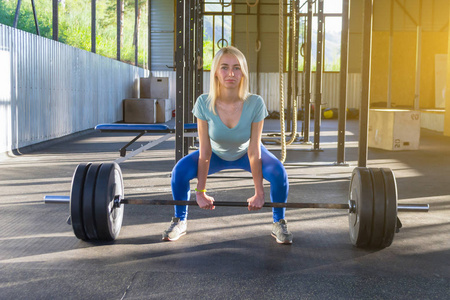 金发女郎做的 deadlift, 就在镜头前, 在健身房的背景下, 在体育设备和绿色山脉在阳光照射下, 在体育馆里拿着举重的杠铃