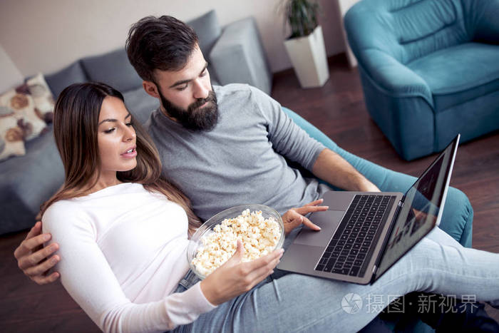快乐的年轻夫妇坐在沙发上的家与爆米花和看电影的笔记本电脑