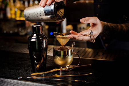 酒保与纹身倒入新鲜可口的夏日饮料从振动筛到酒吧柜台上的老式铜杯