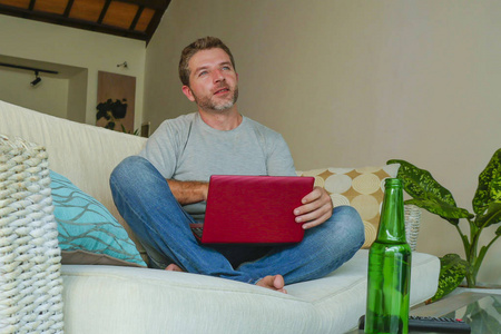生活在室内的年轻迷人的和英俊的快乐男人坐在家里沙发沙发上工作的生活方式与笔记本电脑使用上网本轻松舒适的啤酒瓶