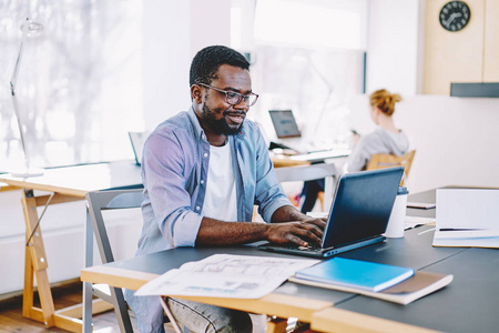 微笑的非洲裔美国男学生打字膝上型电脑在社交网络与朋友聊天在图书馆, 积极的深色皮肤自由职业者满意网上业务通过上网本
