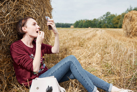 女孩坐在干草堆附近, 用口红涂嘴唇。