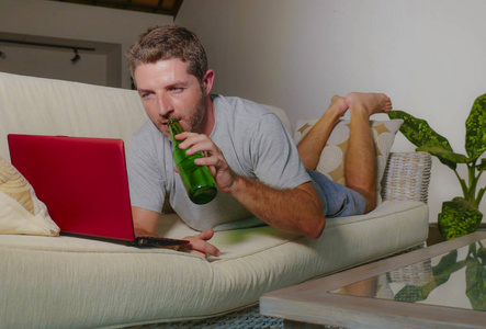 生活在室内的年轻有魅力和快乐的男人的肖像躺在家里沙发沙发上轻松使用互联网上的笔记本电脑看在线电影或远程工作和喝啤酒瓶