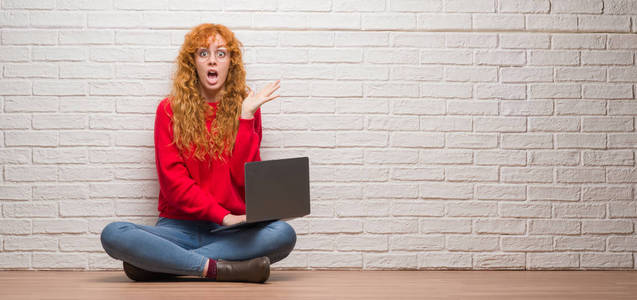 年轻红头发的女人坐在砖墙上使用电脑笔记本非常高兴和兴奋, 优胜者表达庆祝胜利尖叫与大微笑和举手