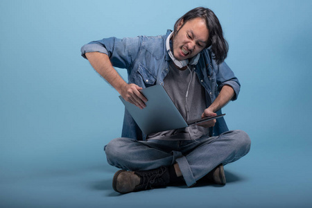 年轻的大胡子男子坐在地板上使用计算机在蓝色背景。非常愤怒的亚洲年轻的时髦使用笔记本电脑, 全身射击。年轻一代时髦的工作理念