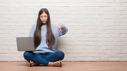 年轻的中国妇女坐在地板上墙上使用膝上型电脑与愤怒的脸, 消极的迹象显示不喜欢拇指向下, 拒绝概念