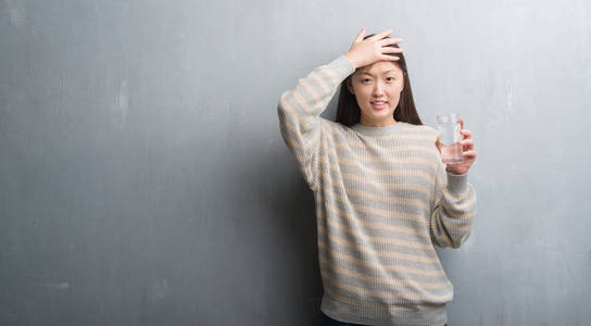 年轻的中国女人在灰色的墙上喝着水, 用手在头上, 感到羞愧和惊讶的脸, 愤怒和沮丧。对错误的恐惧和不安