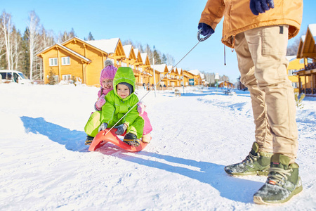 父亲拉雪橇与快乐的小男孩和女孩沿雪路上在美丽的冬季下午度假胜地地区