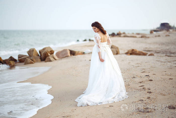 一个穿着白色婚纱的漂亮新娘正沿着海滩散步。波浪靠近她的衣服。美丽的日落气氛, 夏日的傍晚。婚礼理念