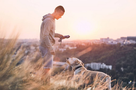 早上和狗一起散步。年轻男子和他的拉布拉多猎犬在草地上对城市的日出