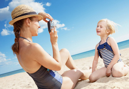 微笑的年轻母亲和孩子泳装拍照与复古电影照片相机在海滩上