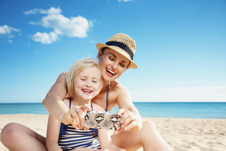 快乐的年轻母亲和女儿泳装采取自拍与智能手机在海滩上