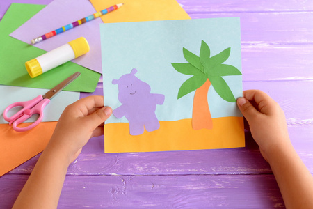 儿童持有纸卡与河马和棕榈树。彩色纸，剪刀，铅笔，胶水粘丁香木背景上。学龄前儿童的的可爱且容易手工工艺品