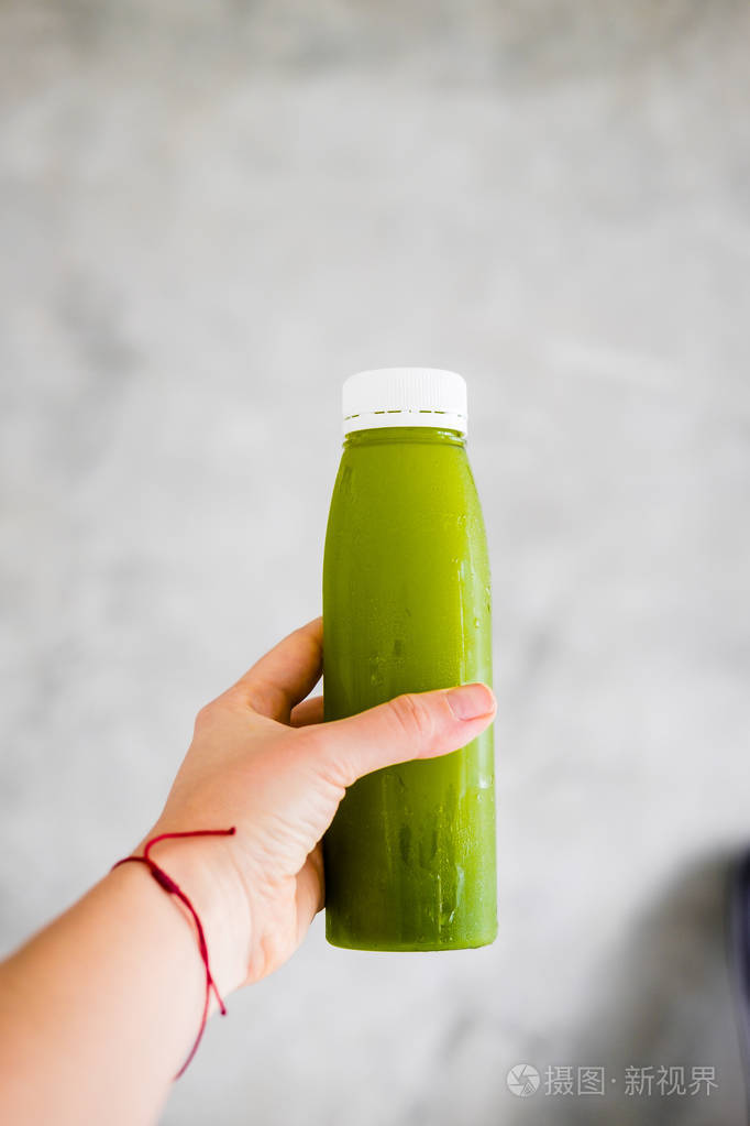女人的手拿着一瓶冷榨的绿色果汁对抗灰墙.