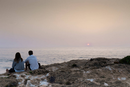 一对年轻夫妇看着太阳设置在地平线上, 而在靠近悬崖在蒙特 Clerigo, 葡萄牙