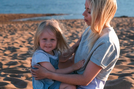 美丽的金发碧眼的母亲和女儿在海滩上拥抱日落