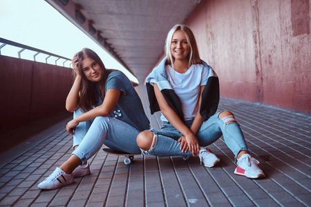两个美丽的时髦女孩坐在滑板上的人行道下桥