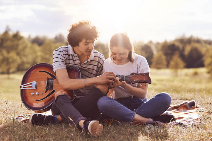 可爱的情侣坐在绿草的腿上, 弹吉他, 唱歌, 享受团结。可爱的年轻女性学会如何产生声音在乐器, 收到的建议从男朋友