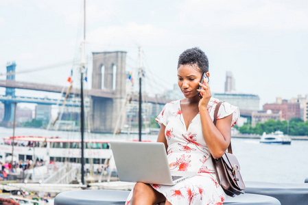 非洲裔美国妇女, 短非洲头发在纽约旅行, 坐在东河边, 电话交谈, 同时在笔记本电脑上工作。曼哈顿, 布鲁克林大桥, 背景船