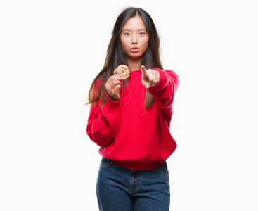 年轻的亚洲妇女吃巧克力片饼干在孤立的背景下指向相机和你, 手势, 正面和自信的手势从前面