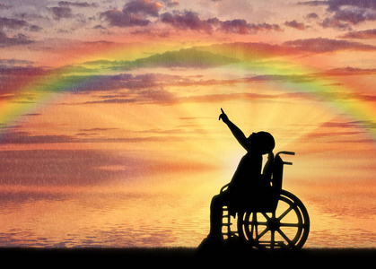 一个残疾儿童女孩的剪影, 坐在轮椅上, 在彩虹的背景下显示一根手指。残疾儿童生活的概念性形象