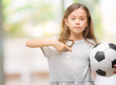 黑头发的西班牙女孩拿着足球球与惊讶的脸指向自己的手指