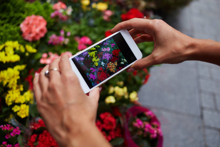 Instagram 摄影师博客研讨会的概念。关闭女性手拿着电话和拍照的时尚花。街头集市上五颜六色的花朵。文本的空间。选择性聚焦