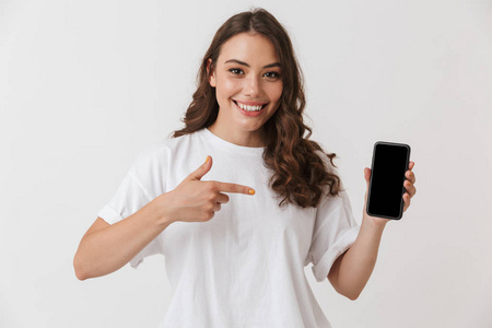 一个微笑的年轻的休闲黑发女人的肖像指向在空白屏幕上的手机在白色背景下隔离