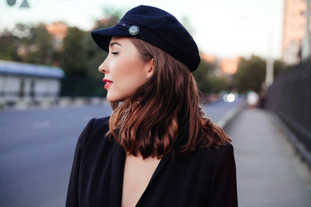 有魅力的现代女性站在城市街道上。时尚的年轻女子走在街上的城市穿着黑色的帽子和夹克。时尚夏日照片, 旧城背景。望向侧面
