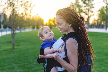 年轻快乐的母亲与婴孩在因此背包旅行一起。阳光明媚的夏日在公园里。现代父母的观念