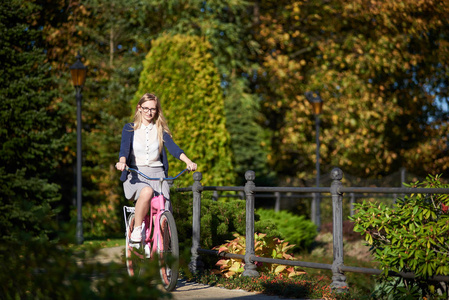 愉快的金发长发美女在眼镜和休闲服装骑自行车现代粉红色女士自行车沿狭窄的公园桥梁在散景背景美丽的不同的绿树