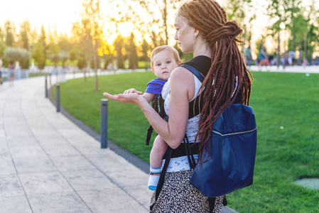年轻快乐的母亲与婴孩在因此背包在公园散步。阳光明媚的夏日。现代父母的观念