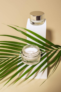 在米色背景上的白色立方体上的玻璃罐子上的绿色棕榈叶和体霜的特写视图