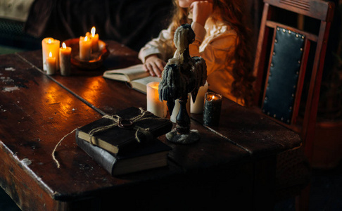 老式的木桌上有蜡烛的烛台。一个红头发的女孩穿着老式的衣服坐在桌子旁, 在烛光下看着一本旧书在木桌上。