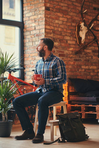 穿着蓝色羊毛衬衫和牛仔裤的英俊胡子的时髦男, 坐在阁楼内部的工作室的木凳上, 看着远处的一杯咖啡。