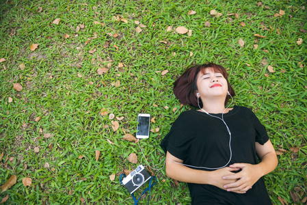 年轻的亚洲妇女躺在绿色的草地听音乐在公园与寒意的情绪。在草地上放松的年轻女子与她的音乐播放列表。户外活动在公园概念