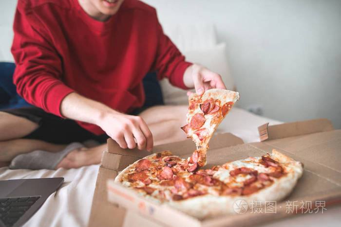 穿着红色运动衫的年轻人坐在家里的床上, 从盒子里拿出一件开胃的比萨饼。男人的手拿一块美味的比萨饼。这个十几岁的孩子要吃一块比萨饼