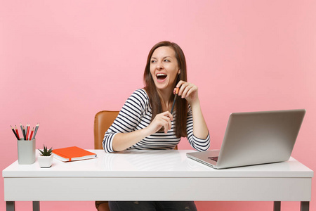 年轻开朗的女人与张开的嘴拿着铅笔看着一旁坐在白色办公桌上的工作与当代 pc 笔记本电脑在柔和的粉红色背景隔离。成就事业的事业理念