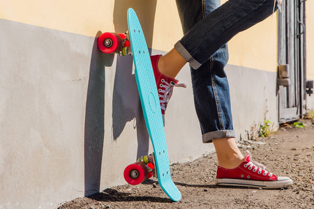 在红色运动鞋和蓝色便士滑板的女孩关闭脚, 粉红色的车轮站在墙边。都市风光, 城市生活。运动, 健身生活方式