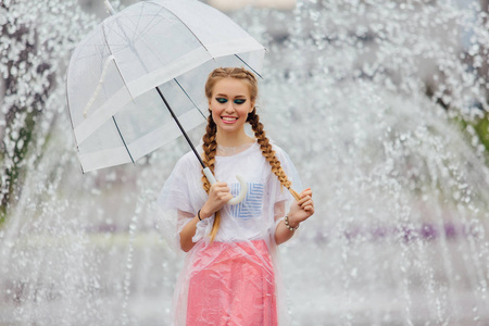 年轻漂亮的女孩与两个辫子在黄色靴子和透明的雨伞站在喷泉附近。城市的雨天